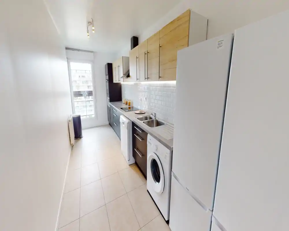 Cuisine moderne en longueur, lumineuse, avec placards hauts en bois, meubles bas foncés, appareils intégrés et évier double. L'espace est propre, bien éclairé et adapté aux annonces locatives.