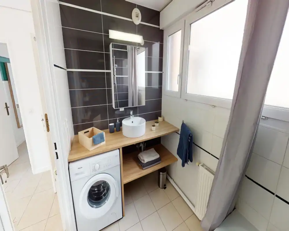 Salle de bain moderne et lumineuse avec vasque ronde sur plan, miroir mural et grandes fenêtres dépoli ; un lave-linge est intégré sous le plan en bois.