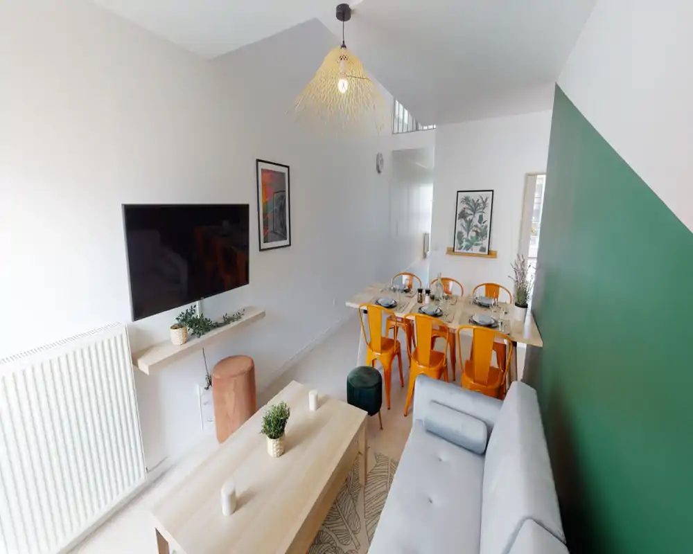 Pièce de vie lumineuse et contemporaine avec un canapé gris clair, télévision murale, table basse en bois et une table à manger avec six chaises orange. Décoration moderne, plantes et textiles apportent une atmosphère chaleureuse et soignée.