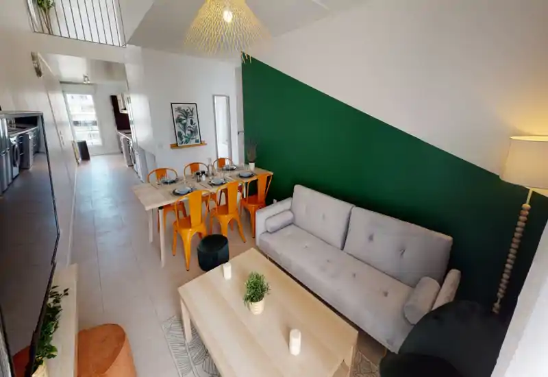 Salon moderne et lumineux avec canapé gris confortable, table basse en bois et coin repas avec chaises orange devant un mur d'accent vert.