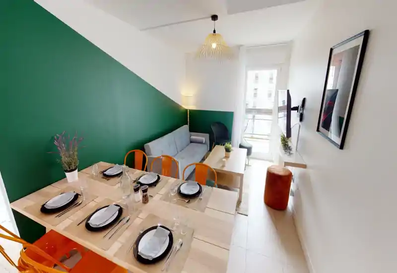 Espace séjour et salle à manger élégant avec mur vert, canapé confortable, table dressée pour six et belle lumière naturelle depuis la porte-fenêtre — idéal pour recevoir.