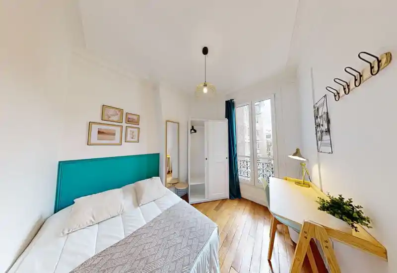 Camera da letto luminosa e accogliente con testiera turchese, letto comodo, pavimento in legno e una scrivania vicino alla finestra — perfetta per lavoro e relax.