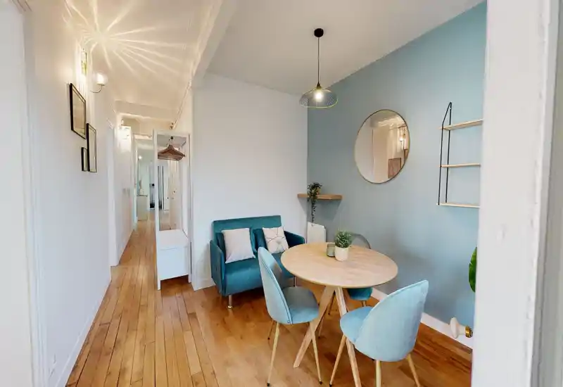 Accogliente angolo pranzo luminoso con un piccolo tavolo rotondo in legno e quattro sedie in velluto davanti a una parete accentata azzurra — ideale per pasti o colazioni.