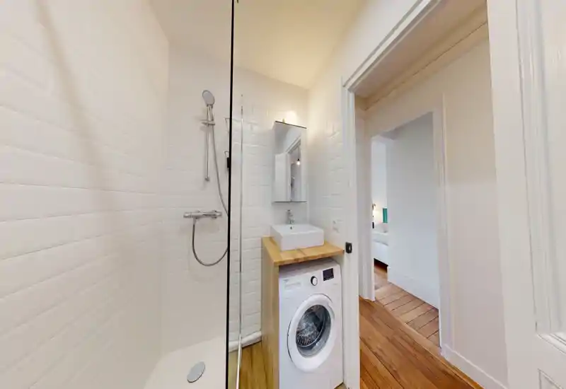 Bagno compatto e moderno con doccia walk-in, piastrelle bianche effetto subway, un piccolo mobile con lavabo e una lavatrice a carica frontale.