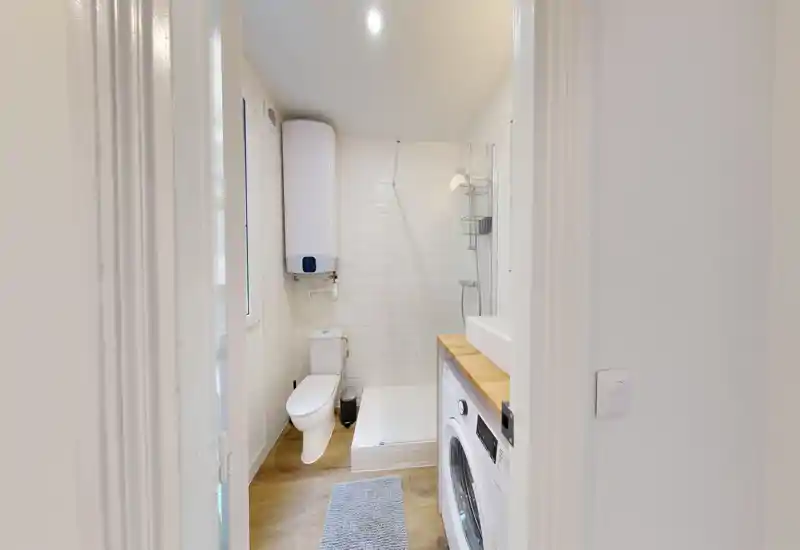 Bagno moderno e luminoso con doccia walk-in, wc e lavatrice sotto un piano in legno. Piastrelle bianche e pavimento chiaro donano un aspetto pulito e arioso.