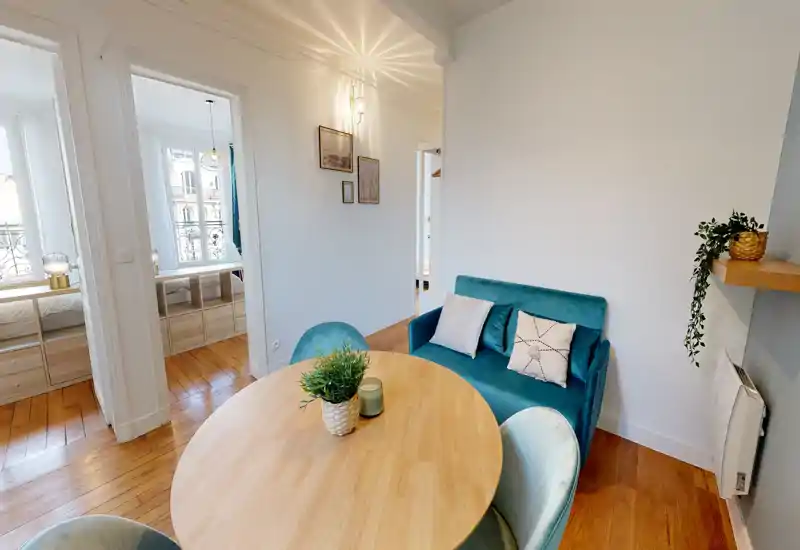 Accogliente angolo pranzo soleggiato con tavolo rotondo in legno, sedie in velluto e un piccolo divano turchese, luce naturale e piante decorative.