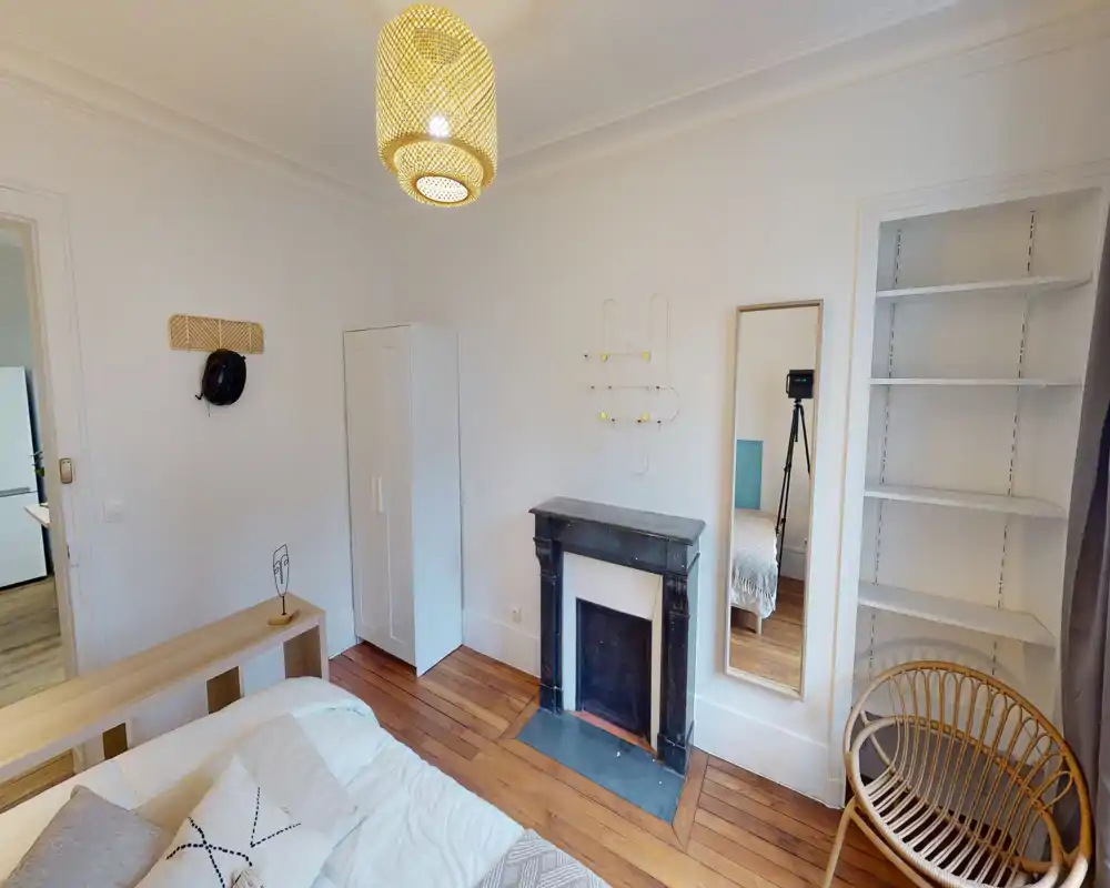 Chambre lumineuse et chaleureuse avec parquet, lit partiellement visible, armoire et cheminée décorative ; style moderne et épuré, grand miroir ajoutant de la profondeur.