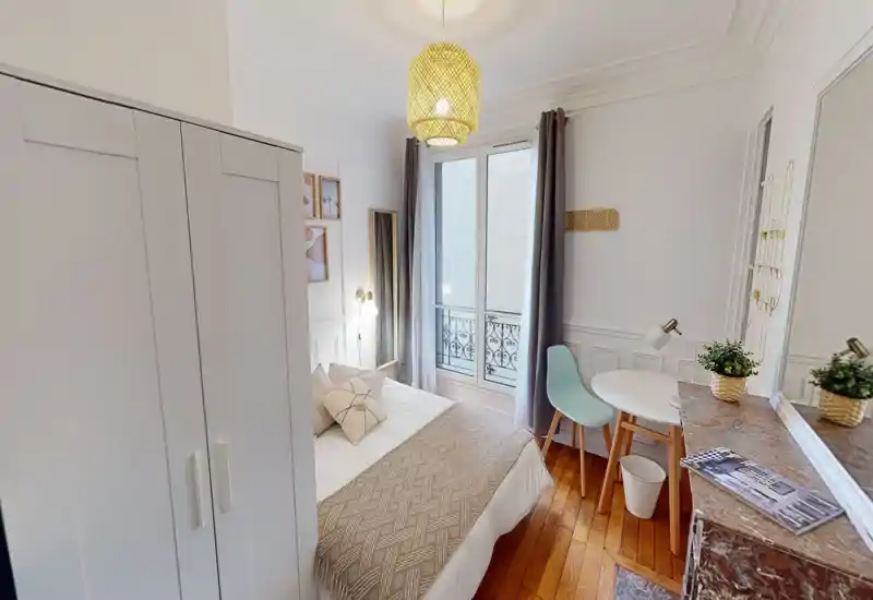 Chambre lumineuse et chaleureuse avec un grand lit bien fait, coussins décoratifs et suspension chaleureuse. Grande fenêtre avec rideaux pour la lumière naturelle et petite table avec chaise pour un espace de travail pratique.