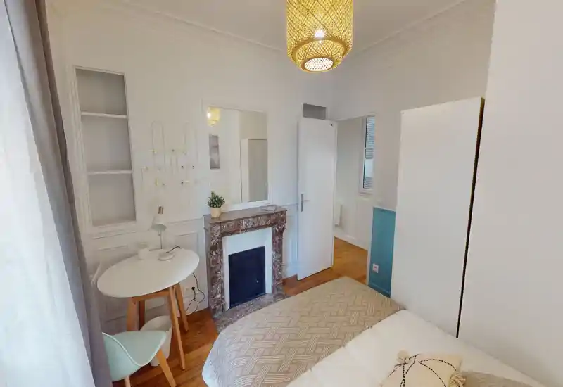 Chambre lumineuse et élégante avec lit visible, jeté à motif, cheminée décorative et petit espace bureau près de la fenêtre — parfaite pour un séjour citadin cosy.