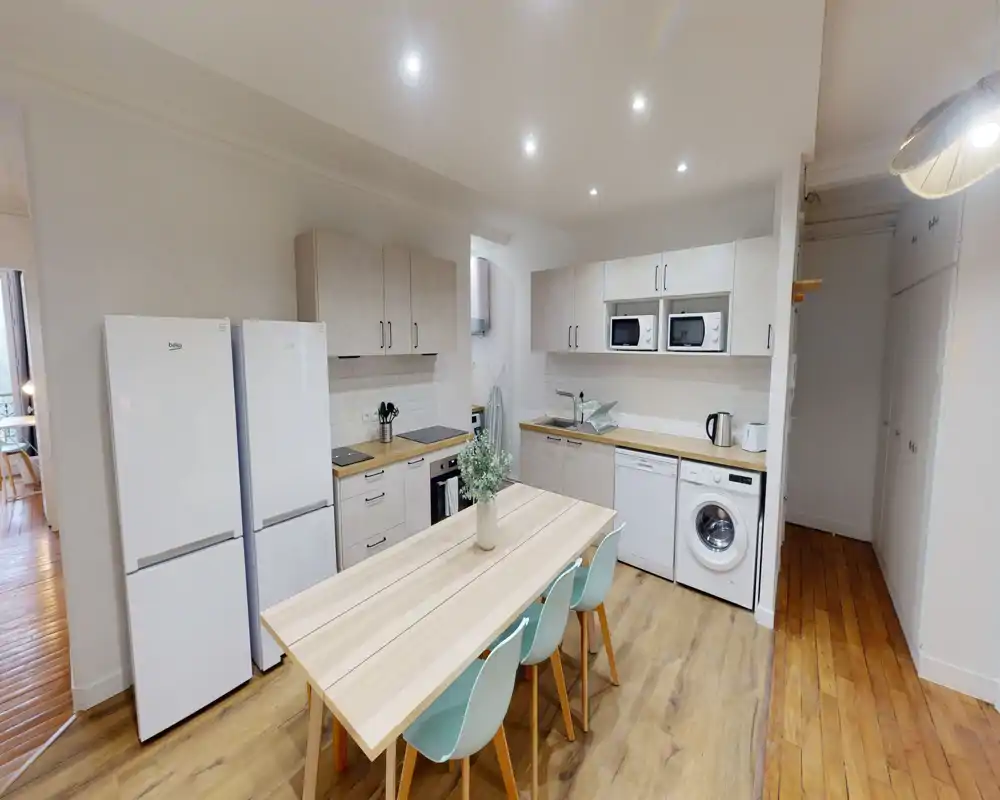 Cuisine moderne équipée avec électroménagers intégrés, table en bois et quatre chaises pastel ; surfaces claires et éclairage encastré apportent une atmosphère lumineuse et fonctionnelle.