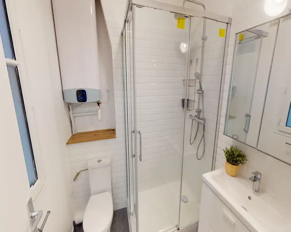 Salle de bain moderne et compacte avec cabine de douche vitrée, robinetterie murale, lavabo avec meuble et miroir, et WC ; carrelage blanc lumineux et équipements propres.