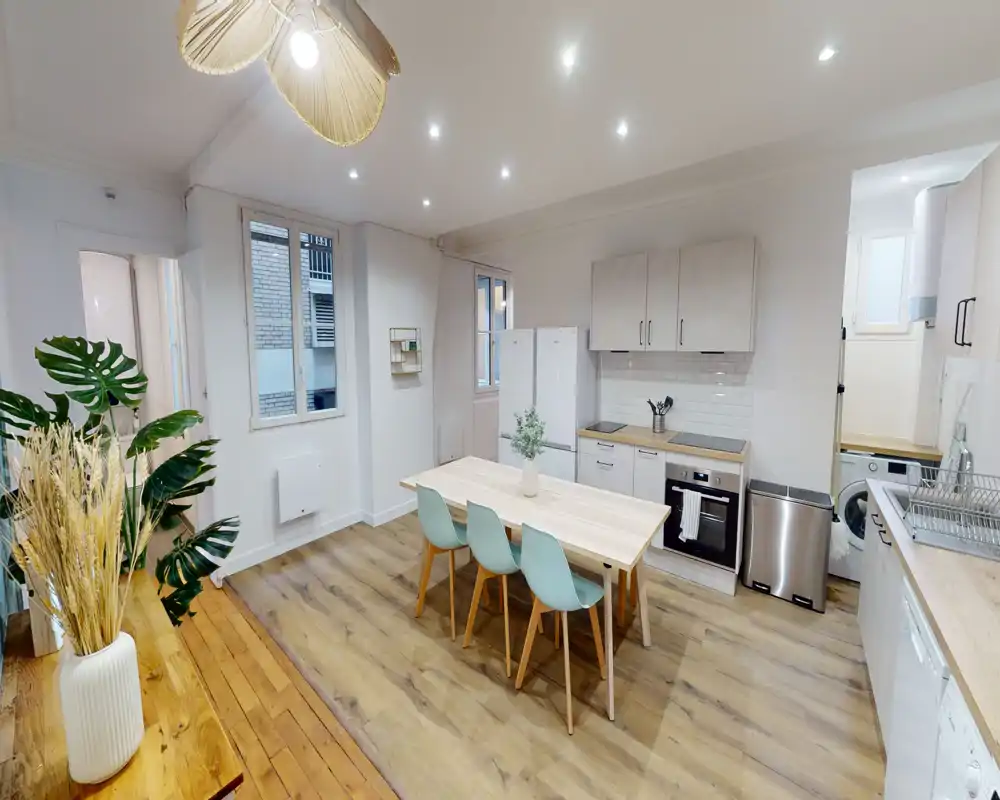 Cuisine moderne et lumineuse avec four intégré, réfrigérateur et une table centrale en bois avec quatre chaises. Parquet clair, meubles blancs et plantes décoratives créent un espace chaleureux et accueillant, idéal pour les repas et les moments conviviaux.