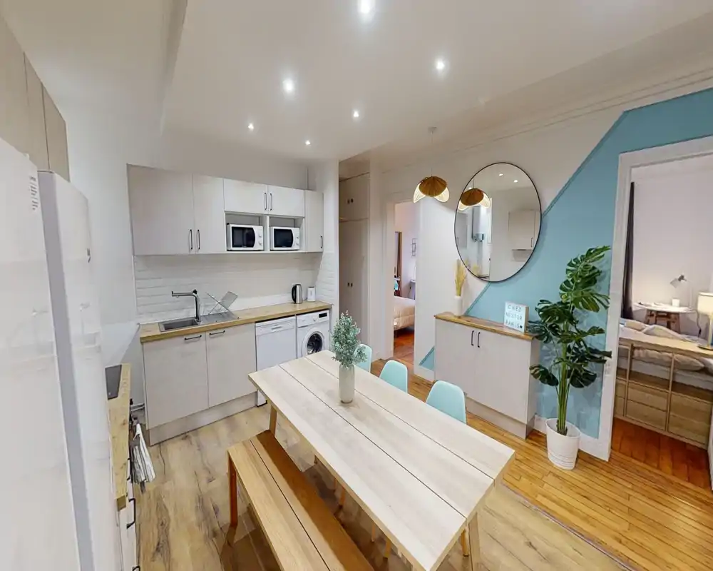 Espace repas en open-space avec table en bois clair et banc, adjacent à une cuisine compacte. L'espace est décoré avec des chaises pastel, une plante et un miroir rond, offrant un coin repas lumineux et accueillant.