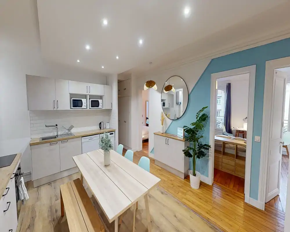 Cuisine ouverte lumineuse avec table à manger en bois et banc, meubles blancs modernes, micro-ondes et lave-vaisselle intégrés, et plantes décoratives pour une ambiance accueillante.