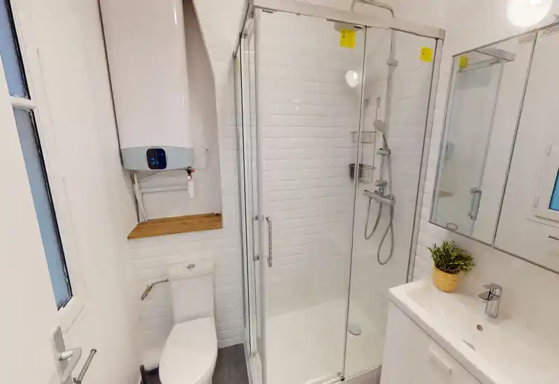 Salle de bain moderne et compacte avec cabine de douche vitrée, robinetterie murale, lavabo avec meuble et miroir, et WC ; carrelage blanc lumineux et équipements propres.