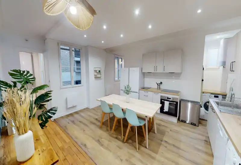 Cuisine moderne et lumineuse avec four intégré, réfrigérateur et une table centrale en bois avec quatre chaises. Parquet clair, meubles blancs et plantes décoratives créent un espace chaleureux et accueillant, idéal pour les repas et les moments conviviaux.