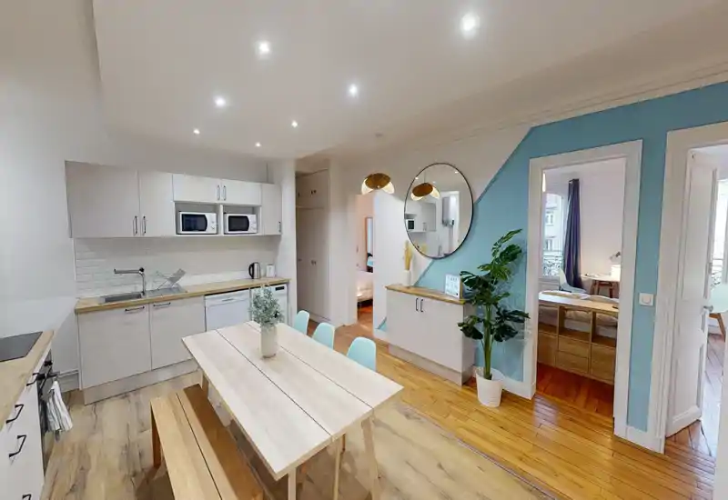 Cuisine ouverte lumineuse avec table à manger en bois et banc, meubles blancs modernes, micro-ondes et lave-vaisselle intégrés, et plantes décoratives pour une ambiance accueillante.