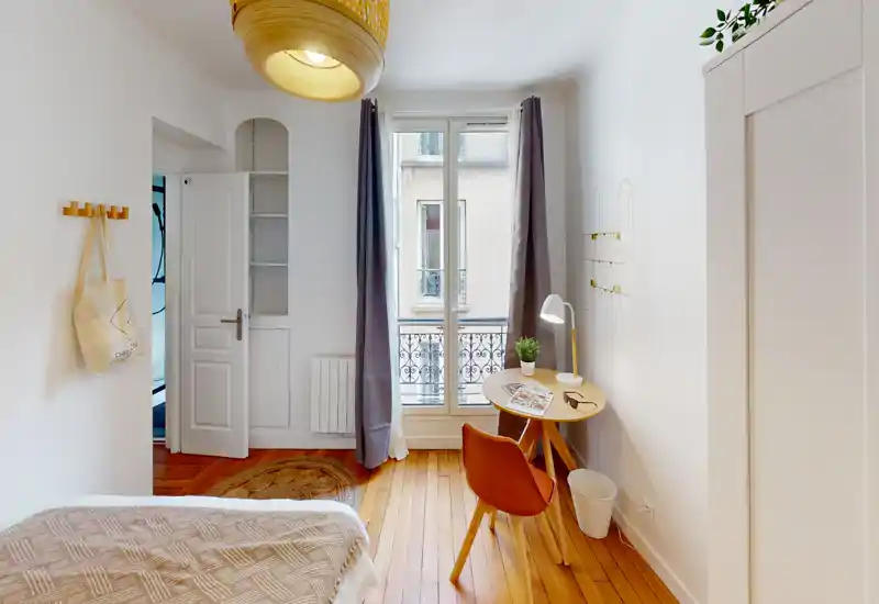 Chambre simple lumineuse et chaleureuse avec parquet, lit soigné, petit bureau près de la fenêtre et lumière naturelle douce — idéale pour un séjour confortable.