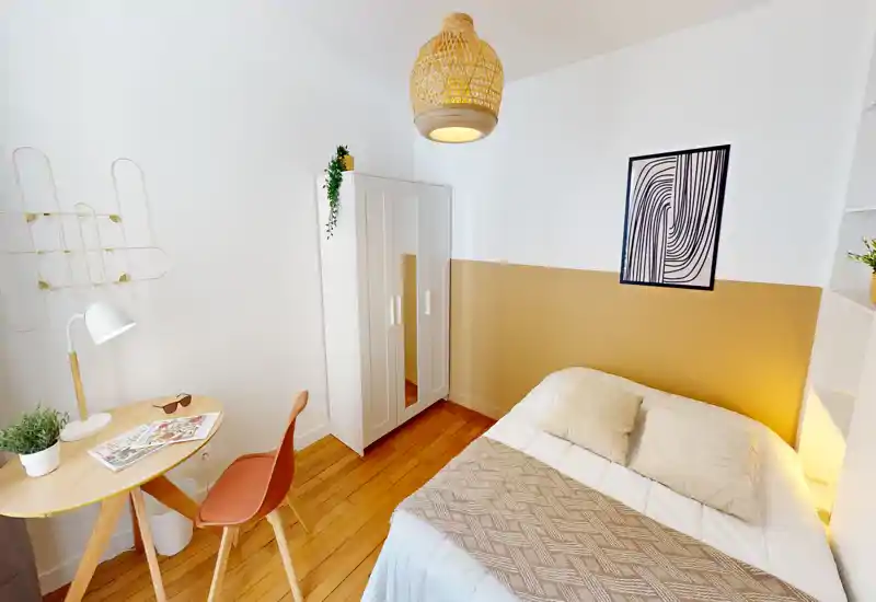 Chambre chaleureuse et lumineuse avec un lit double en linge neutre, suspension en osier, petite armoire et bureau compact — idéale pour une personne seule ou un couple recherchant un espace cosy et stylé.