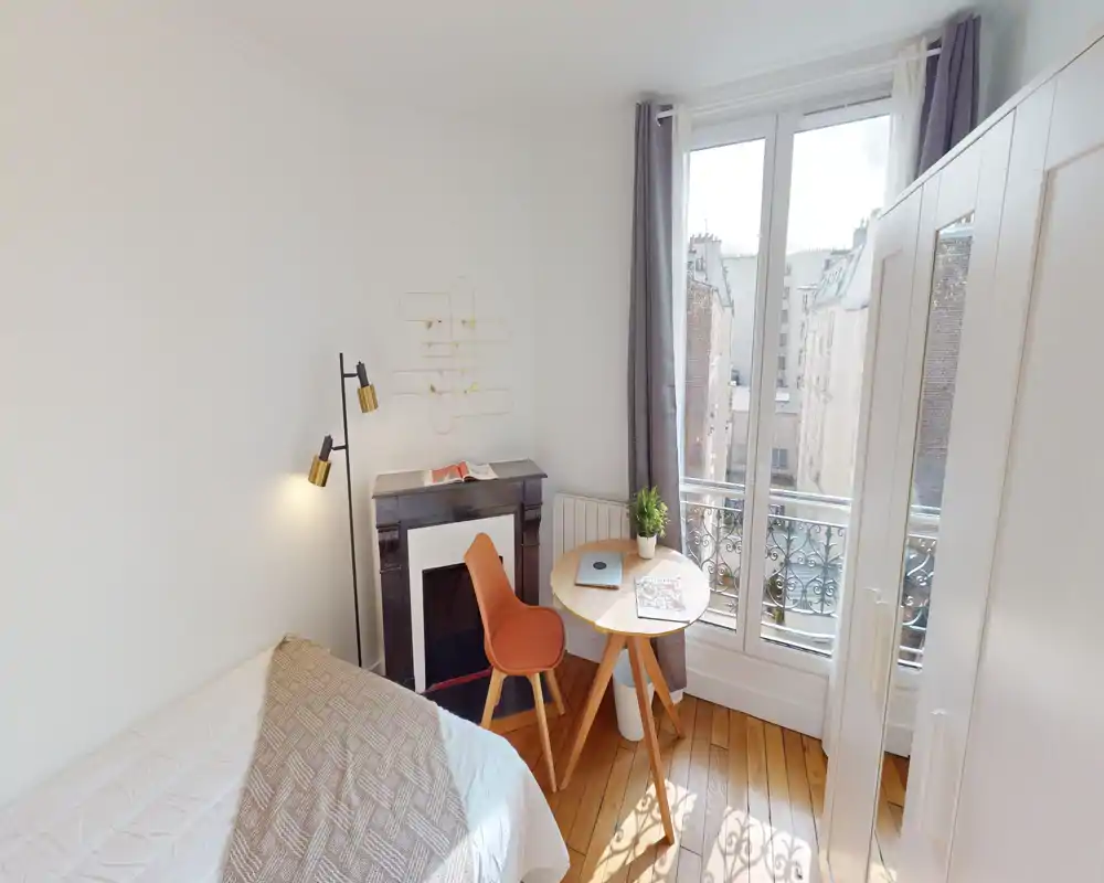 Camera da letto luminosa e compatta con letto singolo, pavimento in legno e un piccolo tavolo rotondo vicino alla finestra. Spazio moderno e arioso con luce naturale e arredi essenziali.