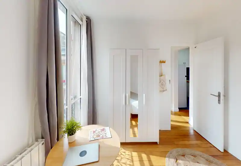Angolo camera luminoso e minimale con pavimento in legno, armadio con ante a specchio e scorcio del letto attraverso la porta. Ampia finestra con tende che lascia entrare molta luce naturale; una piccola scrivania con pianta aggiunge un tocco accogliente e funzionale.