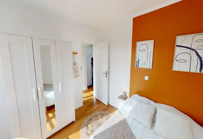 Camera da letto luminosa e accogliente con letto bianco, pavimento in legno naturale e una parete arancione con quadri. Tessuti caldi e un armadio con specchio rendono la stanza piacevole e ben illuminata.