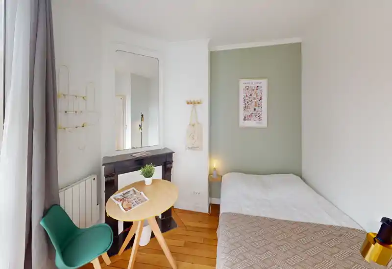 Compact, stylish bedroom with a single bed against a soft green accent wall, wooden floor and a small round table by the window. Natural light and tasteful decor create a bright, cozy atmosphere ideal for short-term rentals.