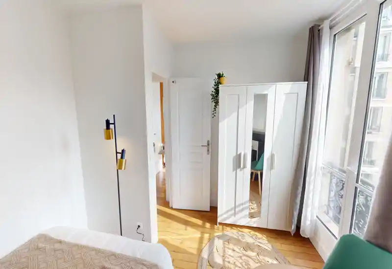 Bright, compact bedroom with a partially visible bed, white wardrobe with mirrored door, wooden floors and large windows that flood the room with natural light.