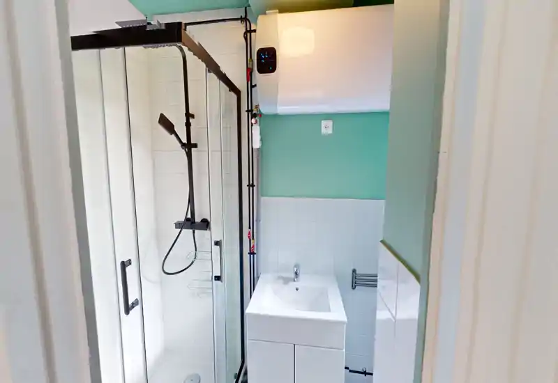 Salle de bains moderne et compacte avec douche vitrée, pommeau de douche mural et petit meuble-lavabo blanc. Carrelage clair et murs vert menthe pour une atmosphère fraîche.