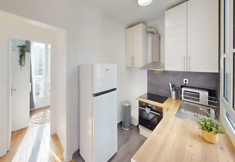 Cuisine moderne compacte avec plan de travail en bois, four et plaque intégrés, évier près de la fenêtre et grand réfrigérateur — lumineuse, bien rangée et adaptée à la vie urbaine.