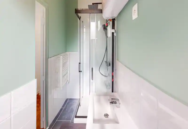 Salle de bain compacte et moderne avec douche vitrée, radiateur mural et lavabo blanc; murs vert menthe et carrelage blanc brillant apportent une impression propre et lumineuse.