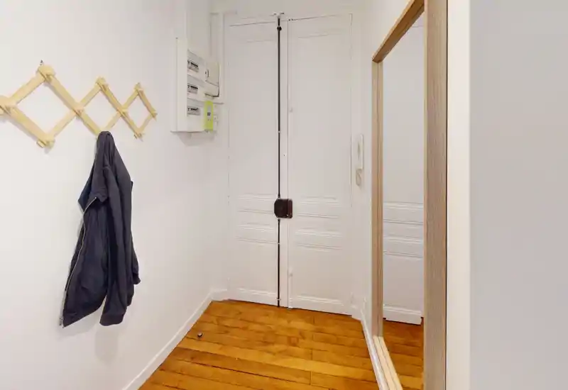 Petit hall d'entrée soigné avec une porte blanche à panneaux, un porte-manteau en bois et un miroir pleine longueur. Le parquet chaleureux apporte du caractère et réfléchit la lumière.