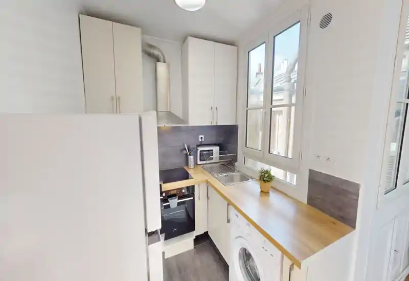 Cuisine compacte et moderne avec placards blancs, plan de travail en bois, four encastré et lave-linge ; lumière abondante par la fenêtre et agencement soigné.