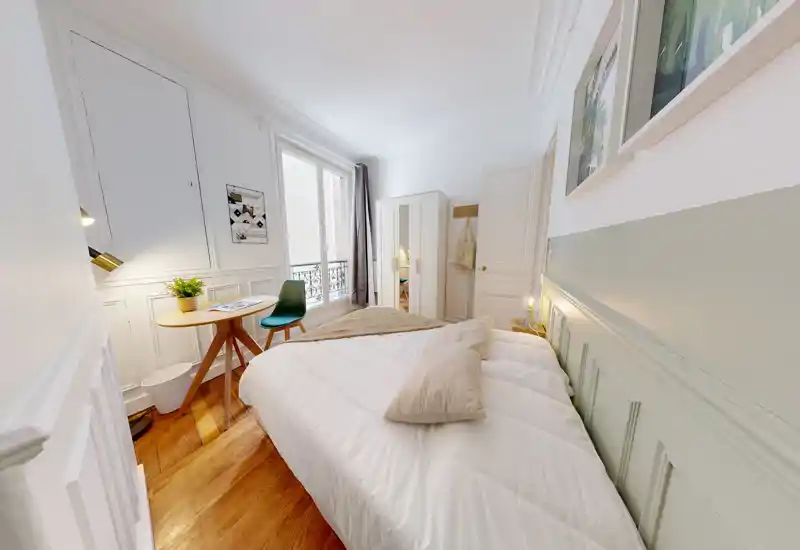 Camera da letto luminosa e accogliente con un ampio letto che domina la stanza, biancheria morbida, pavimenti in legno e un piccolo tavolo vicino alla finestra che garantisce luce naturale e arredo gradevole.