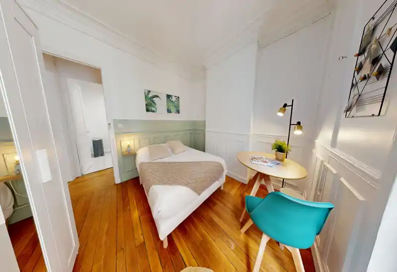 Camera da letto luminosa e arredata con letto matrimoniale, pavimenti in legno, lampade da comodino e un piccolo tavolo rotondo con sedia turchese — ambiente accogliente ideale per soggiorni brevi.