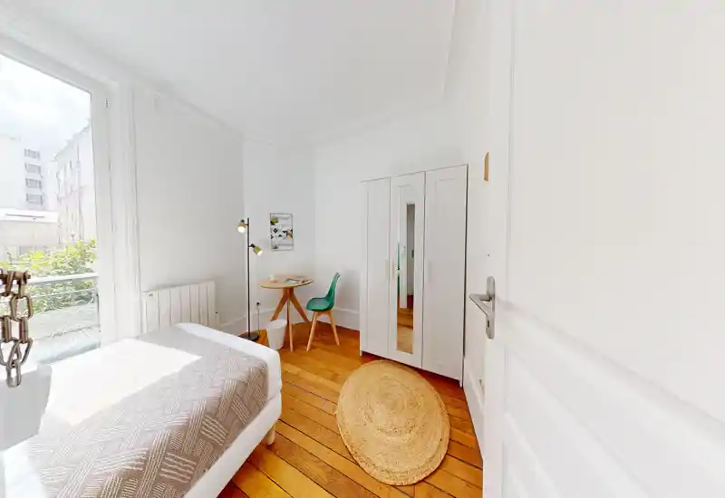 Chambre compacte et lumineuse avec lit confortable, parquet en bois, tapis rond en fibres et petit bureau près de la fenêtre. La grande fenêtre apporte beaucoup de lumière naturelle et la décoration neutre crée une ambiance apaisante.