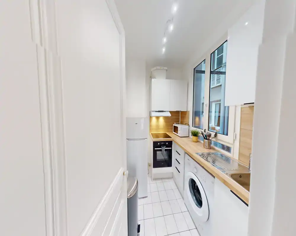 Cuisine en longueur moderne et compacte avec placards blancs, plans de travail en bois, four encastré, lave-linge et une grande fenêtre apportant la lumière naturelle.