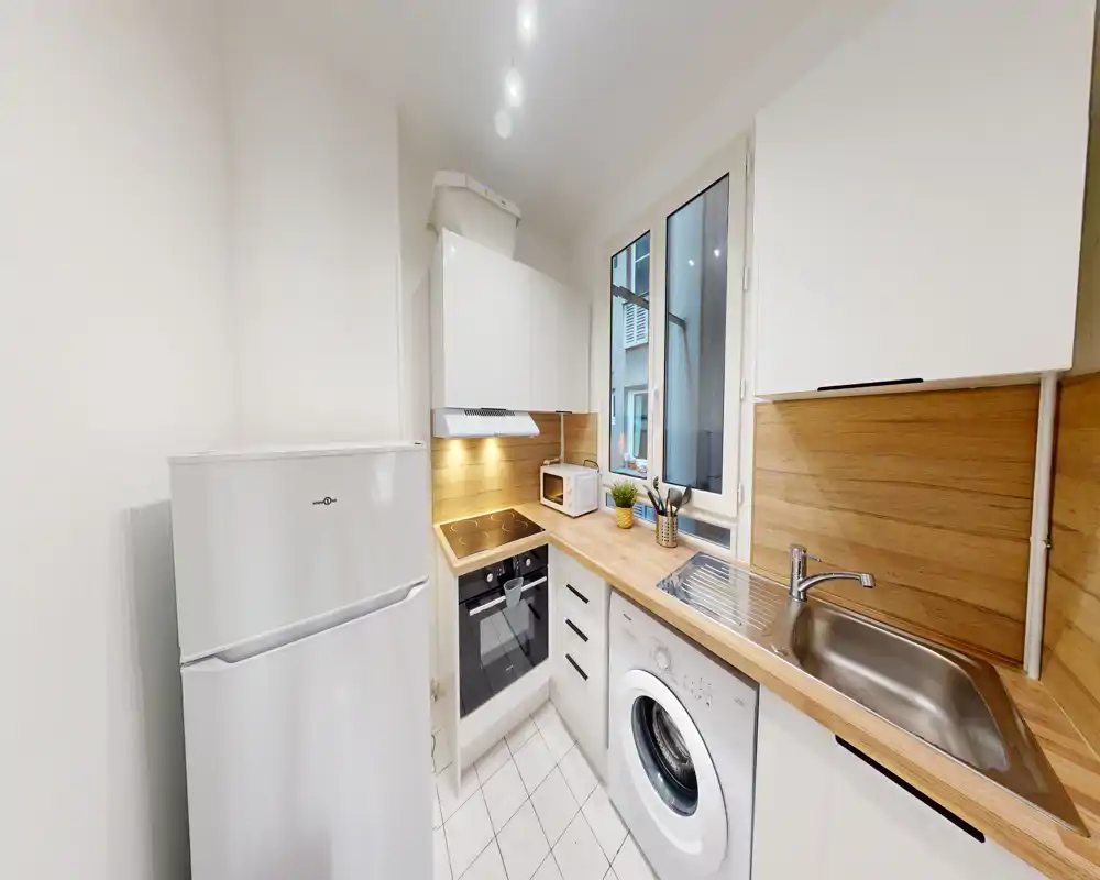Cuisine moderne et compacte avec plan de travail en bois, four et plaque intégrés, évier et lave-linge sous le plan; lumineuse, propre et bien optimisée pour un appartement urbain.