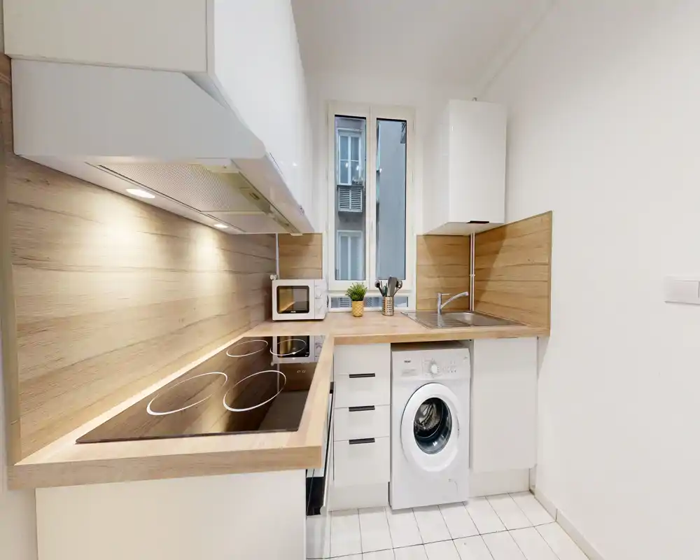 Cuisine moderne et compacte avec plan de travail et crédence effet bois, plaque à induction, évier, micro-ondes et lave-linge intégré ; espace lumineux, propre et adapté aux appartements urbains.