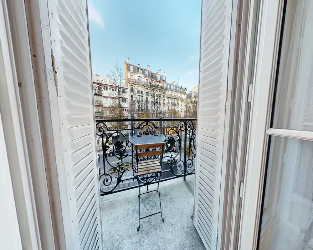 Petit balcon privé avec table bistrot en métal et chaise pliante, garde-corps en fer forgé orné et vue sur des immeubles classiques ; ciel lumineux et lumière naturelle parfaits pour un café le matin.
