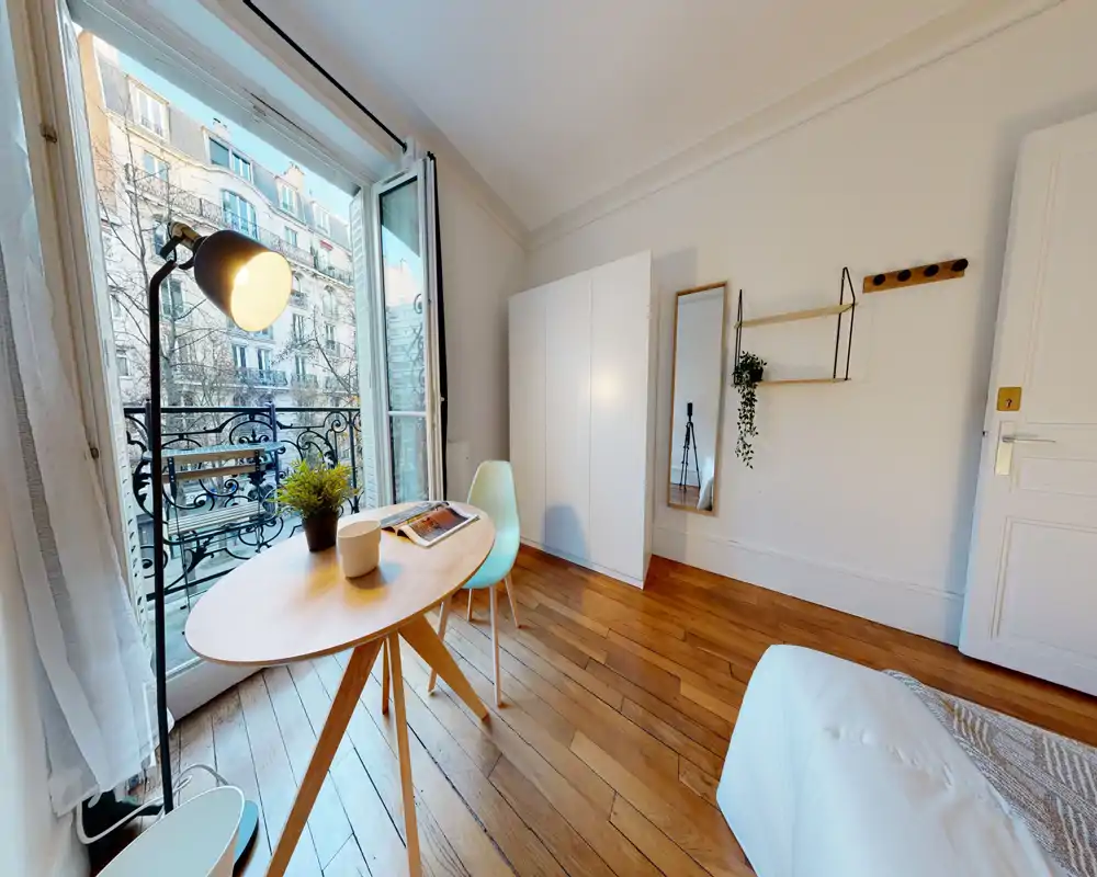 Chambre lumineuse avec parquet, lit compact en bas à droite, petit bureau en bois près d'une fenêtre/balcon et rangements minimalistes — espace clair et soigné.