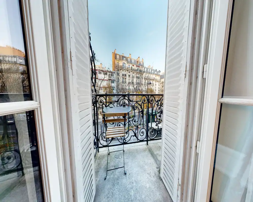 Petit balcon ensoleillé avec balconnet en fer forgé décoratif et une table bistrot compacte avec chaise, offrant une agréable vue sur la ville — idéal pour le café du matin.