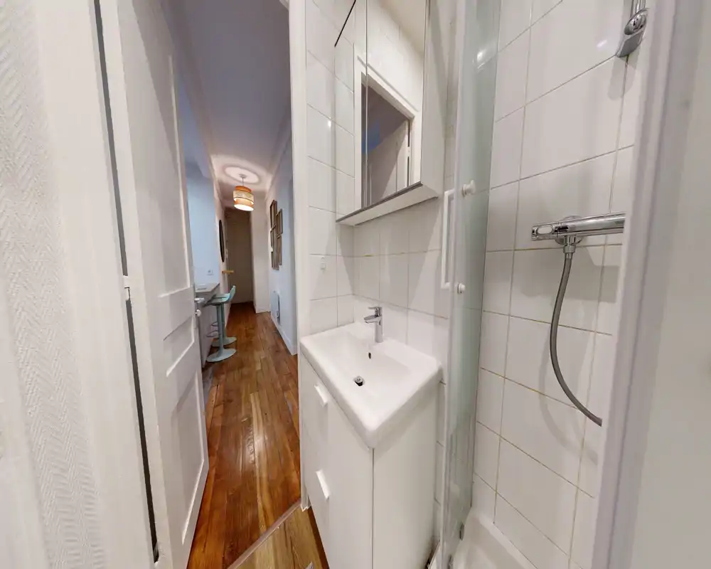 Salle de bain compacte avec douche d'angle à porte vitrée, petit meuble lavabo blanc et armoire-miroir ; carrelage mural blanc et parquet du couloir visible par la porte ouverte.