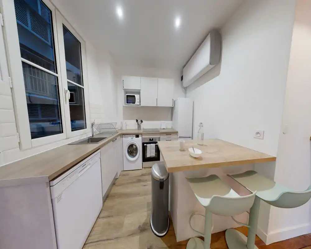 Cuisine moderne et lumineuse avec plans de travail en bois, électroménagers intégrés et un coin petit-déjeuner cosy avec deux tabourets — idéale pour un appartement citadin compact.