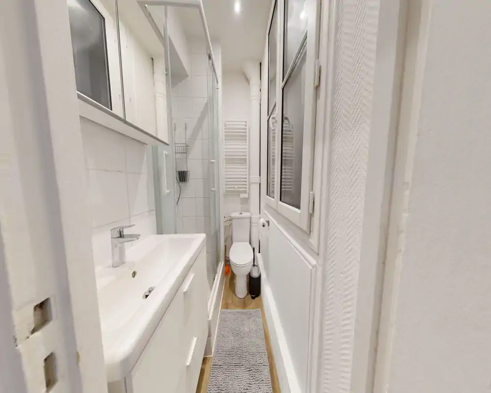 Salle de bains compacte et bien éclairée avec lavabo et meuble à gauche, cabine de douche et WC au fond, radiateur sèche-serviettes et fenêtre sur la droite.