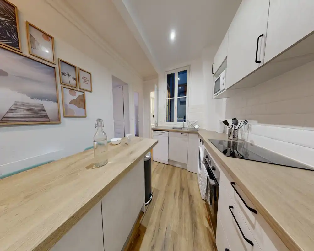 Cuisine moderne et lumineuse avec plans de travail en bois clair, électroménagers intégrés et un îlot central en bois. La décoration chaleureuse et les cadres muraux créent une ambiance accueillante pour cuisiner et prendre des repas rapides.