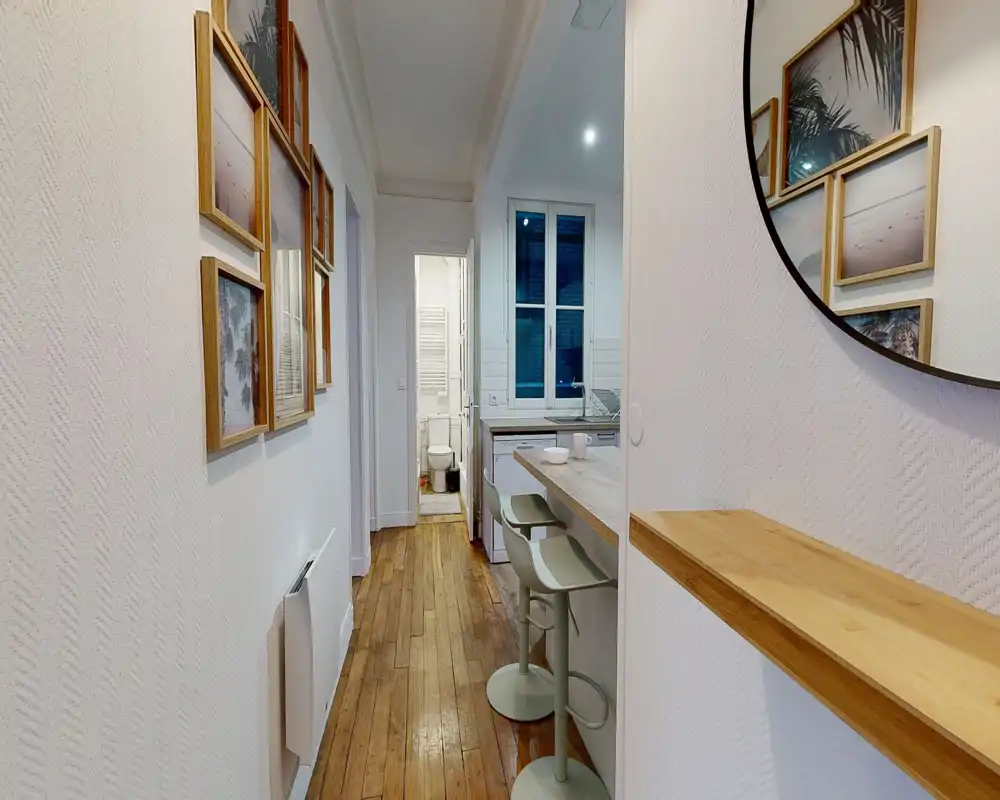 Kitchenette compacte visible au bout d'un couloir étroit avec un bar en bois et deux tabourets, meubles blancs et fenêtre au fond ; le parquet et les cadres aux murs apportent de la chaleur.