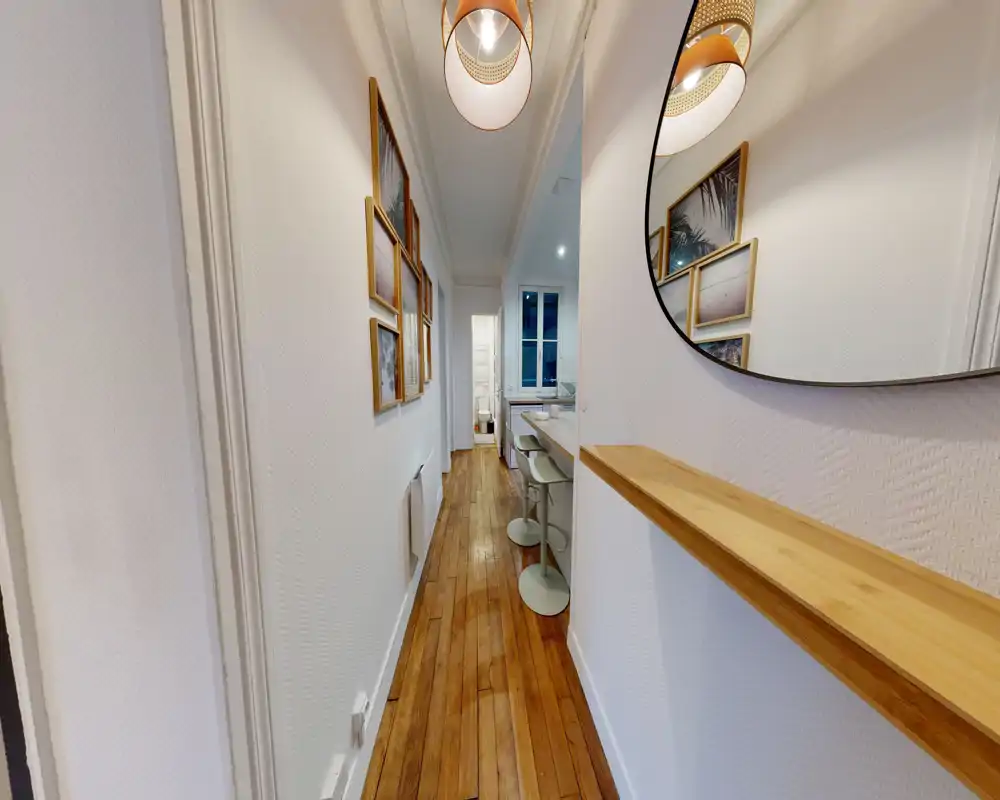 Couloir lumineux et étroit avec parquet, cadres décoratifs et miroir mural ; la perspective mène vers la cuisine avec des tabourets de bar.