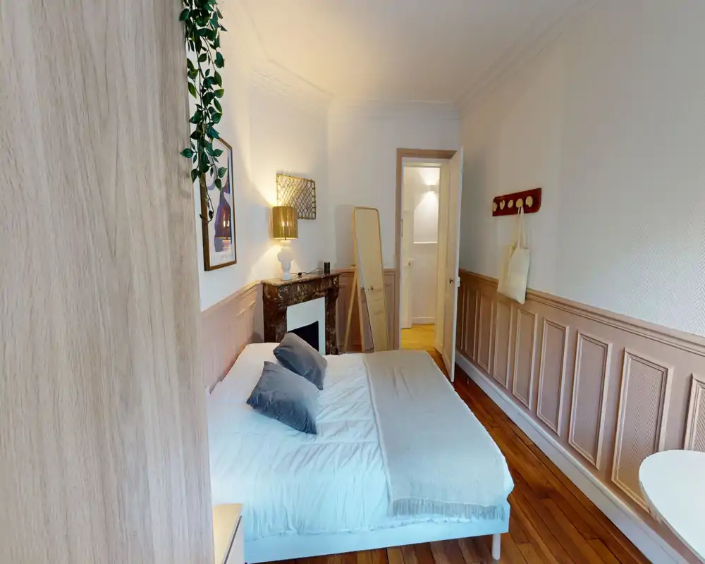 Cozy, well-lit bedroom with a neatly made double bed, decorative pillows, a bedside lamp and classic wood paneling. The room features warm wooden floors and tasteful accents creating a welcoming atmosphere.