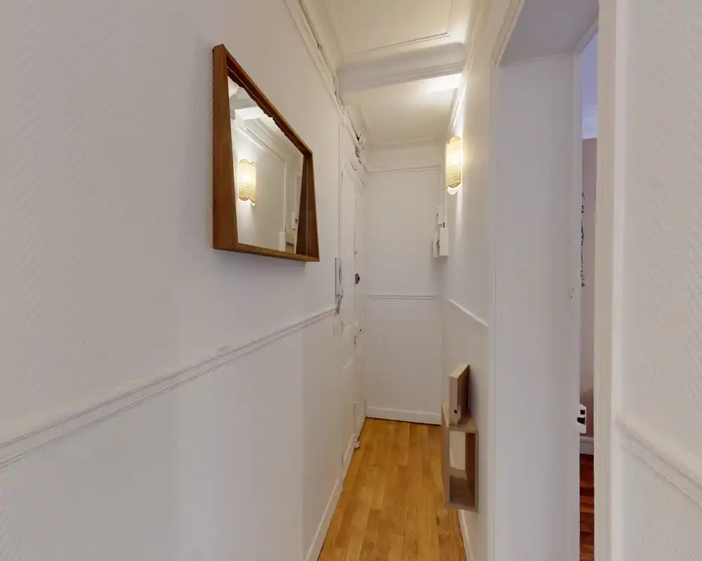 Bright, narrow entrance corridor with white walls, decorative moulding, a wall mirror and warm wooden flooring — practical and welcoming access to the apartment.